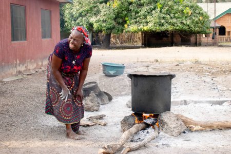 Photo for Opialu, Benue State - March 6, 2021: Smiling Middle Aged African Woman Cooking Using Firewood in a Rural Environment - Royalty Free Image