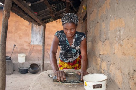 Téléchargez les photos : Opialu, État de Benue - Mars 6, 2021 : Femme africaine utilisant ses mains sur un broyeur local (pierre) pour écraser Melon près de sa maison de boue. - en image libre de droit