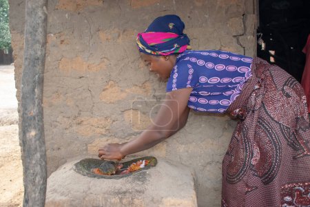 Photo for Opialu, Benue State - March 6, 2021: African Woman using her hands on a Local Grinding Mill (Stone) to mash Melon near her Mud House. - Royalty Free Image