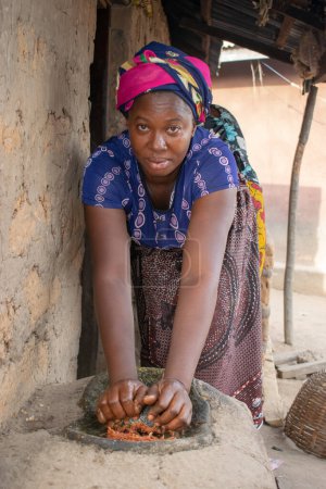 Foto de Opialu, Benue State - 6 de marzo de 2021: Mujer africana usando sus manos en un molino local (piedra) para machacar a Melón cerca de su casa de barro. - Imagen libre de derechos