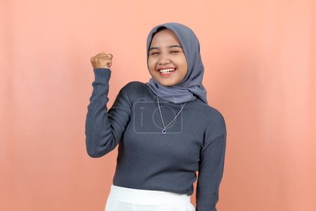 Photo for Excited young Asian woman with clenched fists and raised hands wearing a blue navy sweater isolated peach background - Royalty Free Image