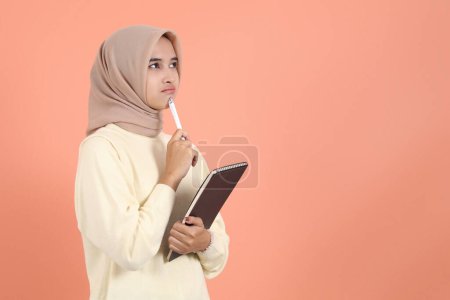 Photo for Pensive young Asian Muslim woman hold book and pen thinking about something - Royalty Free Image