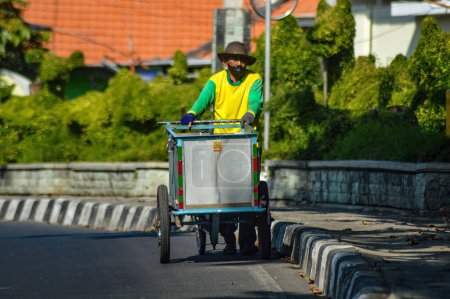 Photo for An old man in yellow clothes pushes an iron cart along the side of a highway in Indonesia, 23 June 2023. - Royalty Free Image
