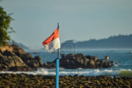 The Indonesian flag fluttering in the wind against the backdrop of a coral rocky beach that looks blurry
