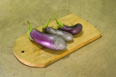 three purple eggplants or Solanum Melongena lying on a cutting board with a plain wooden board background