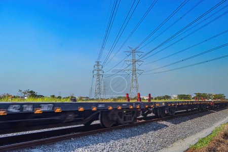 Photo for A series of flat-car trains speeding by against the backdrop of high-voltage power lines during a clear blue sky - Royalty Free Image