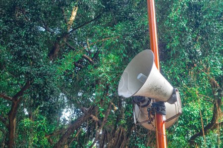 a loudspeaker attached to an iron pole with a backdrop of shady trees