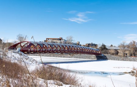 Foto de Un puente peatonal rojo se extiende sobre un río congelado con un cielo azul claro arriba. Las orillas están cubiertas de nieve, y árboles desnudos se pueden ver en la distancia, lo que sugiere un frío día de invierno. - Imagen libre de derechos