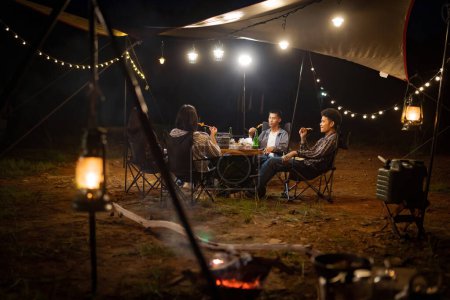 Téléchargez les photos : Jeunes Asiatiques mangeant barbecue sous une toile lors d'une soirée de vacances au lac ayant une tente sous les lumières. - en image libre de droit