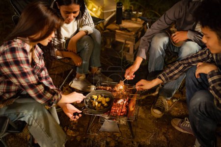 Téléchargez les photos : Jeunes Asiatiques mangeant barbecue sous une toile lors d'une soirée de vacances au lac ayant une tente sous les lumières. - en image libre de droit