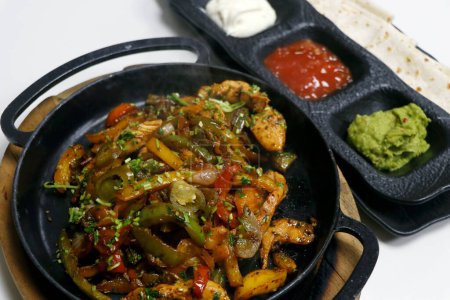 Photo for Mexican food, chicken fajitas serving on a hot smoking sizzling plate with sour cream, guacamole, tomato salsa and tortilla bread - Royalty Free Image