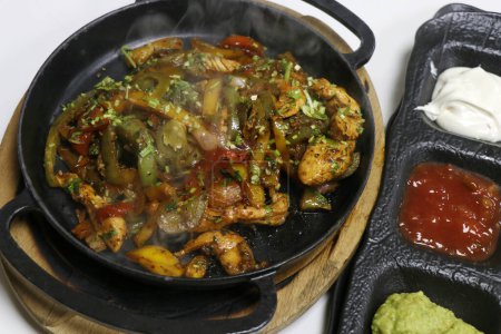 Photo for Mexican food, chicken fajitas serving on a hot smoking sizzling plate with sour cream, avocado guacamole, tomato salsa and tortilla bread - Royalty Free Image