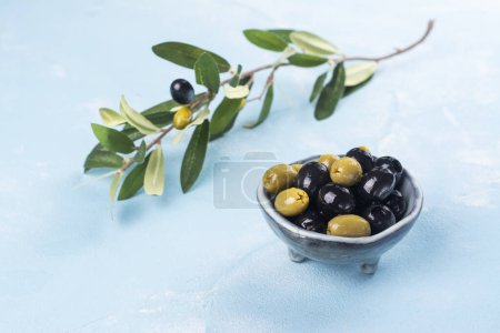 Photo for Black and green greek olives in a bowl on blue sunny background. Copy space - Royalty Free Image