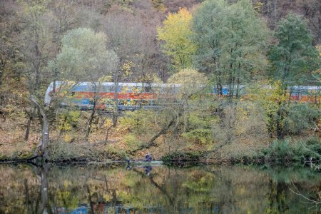 Téléchargez les photos : De l'autre côté de la rivière, un pêcheur pêche. Un train bleu circule sur une voie ferrée. Belle vue sur la rivière d'automne Berounka, saule, forêt et montagnes, Hlasna Treban. tchèque. - en image libre de droit