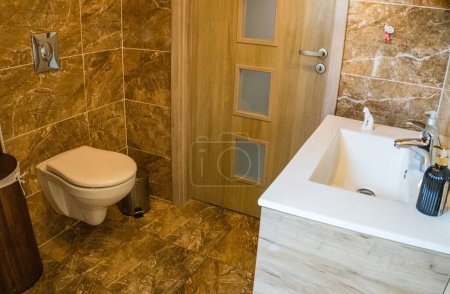 Téléchargez les photos : Salle de bains avec garniture murale en tuiles brunes, panier en bambou et toilettes blanches et lavabo. Photo de haute qualité - en image libre de droit