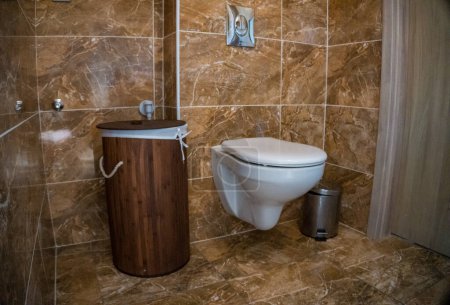 Téléchargez les photos : Salle de bains avec garniture murale en tuiles brunes, panier en bambou et toilettes blanches. Photo de haute qualité - en image libre de droit