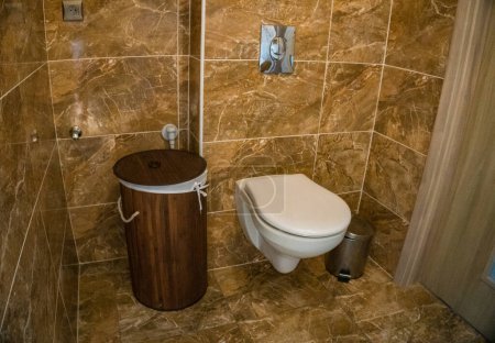 Téléchargez les photos : Salle de bains avec garniture murale en tuiles brunes, panier en bambou et toilettes blanches. Photo de haute qualité - en image libre de droit