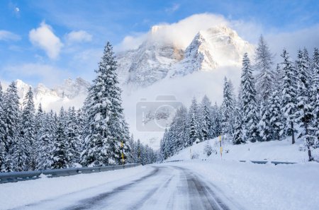Téléchargez les photos : Route glacée traversant une forêt enneigée au pied d'un pic imposant par une journée d'hiver dégagée - en image libre de droit