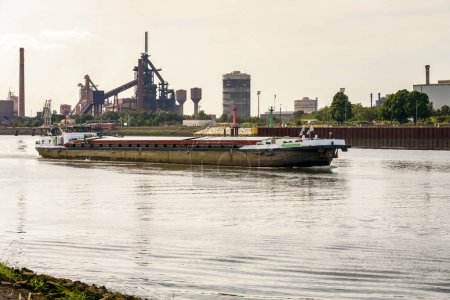 Téléchargez les photos : Barge cargo remontant une rivière au coucher du soleil en été. Une aciérie riveraine est visible en arrière-plan. Brême, Allemagne. - en image libre de droit
