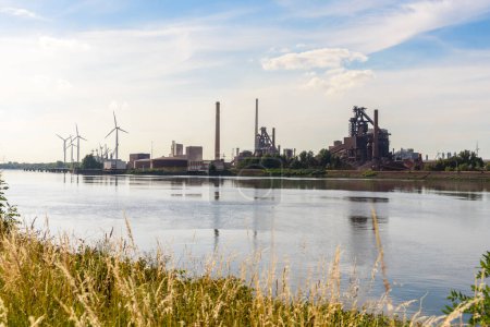 Foto de Gran planta de acero a lo largo de un río en un día soleado de verano. Las turbinas eólicas están en segundo plano. Bremen, Alemania. - Imagen libre de derechos
