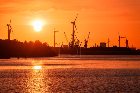 Téléchargez les photos : Bâtiments industriels ainsi que des éoliennes et des grues de quai sur un port fluvial silhouette contre ciel orange au coucher du soleil. Brême, Allemagne. - en image libre de droit