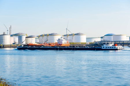 Téléchargez les photos : Barge-citerne naviguant devant un terminal pétrolier avec de grands réservoirs d'acier par une journée d'été claire. Rotterdam, Pays-Bas. - en image libre de droit