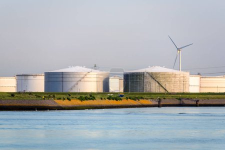 Téléchargez les photos : Grands réservoirs d'huile en acier avec une éolienne en arrière-plan le long d'un port au coucher du soleil. Rotterdam, Pays-Bas. - en image libre de droit