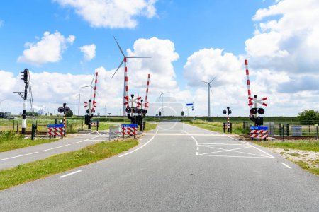 Téléchargez les photos : Level Crossing with Signals and Warning Signs along both a road and a bicycle lane in the countryside of Netherlands. A wind farm is in background. Eemshaven, Netherlands. - en image libre de droit