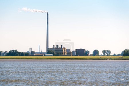 Téléchargez les photos : Chemical plant with a tall smokestack belching out white smoke on the bank of a river on a clear summer day. Nordenham, Germany. - en image libre de droit