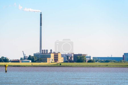 Téléchargez les photos : View of a riverside chemical plant with a tall smokestack belching out white smoke at sunset. Nordenham, Germany. - en image libre de droit