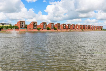 Téléchargez les photos : Rangée de maisons modernes à côté d'un étang dans un ensemble résidentiel par une journée d'été partiellement nuageuse. Groningen, Pays-Bas. - en image libre de droit