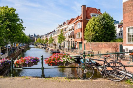 Foto de Canal bordeado de casas de ladrillo tradicionales en un día soleado de verano. Una bicicleta encadenada a una barandilla de puente está en primer plano. La Haya, Países Bajos. - Imagen libre de derechos