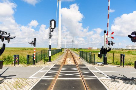 Foto de Cruce a nivel a lo largo de un ferrocarril que conduce a un puerto comercial en un día soleado de verano. Las turbinas eólicas alinean las vías. Eemshaven, Países Bajos. - Imagen libre de derechos