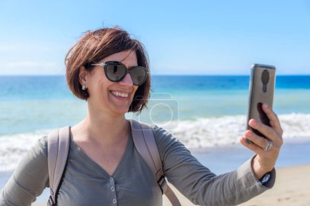Foto de Mujer sonriente tomando una selfie en una playa de arena en California en un día soleado de otoño. Zuma beach, Malibu, CA, Estados Unidos. - Imagen libre de derechos