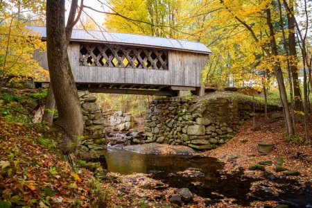 Old pedestrian covered bridge spanning a forest creek in autumn. New Hampshire, USA.