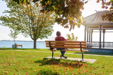 Foto de Mujer disfrutando de una cálida mañana de otoño mientras se sienta en un banco en el parque junto al lago. Kinston, ON, Canadá. - Imagen libre de derechos
