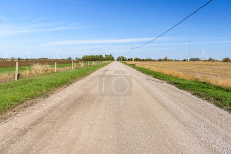 Photo for Straight gravel country road through farmland dotted with wind turbines on a clear autumn day. Wolfe Island, ON, Canada. - Royalty Free Image