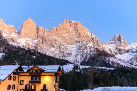 Foto de Edificio de apartamentos tradicional al pie de poderosos picos rocosos nevados que brillan con la luz del atardecer en los Alpes en invierno - Imagen libre de derechos