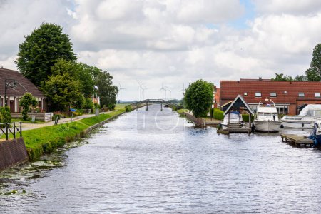 Téléchargez les photos : Canal à travers une petite ville à la campagne des Pays-Bas par une journée d'été nuageuse. Un parc éolien est visible en arrière-plan. - en image libre de droit