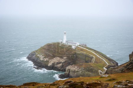 Historischer Leuchtturm auf der Spitze einer kleinen felsigen Insel vor der zerklüfteten Küste an einem nebligen und regnerischen Sommertag. South Stack Leuchtturm, Heilige Insel, Anglesey, Wales, Großbritannien.