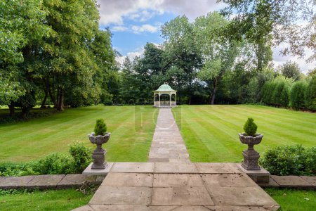 Deserted gazebo at the far end of a stone path through a lawn surrounded by trees in a park on partly cloudy summer day. Aston, England, UK.