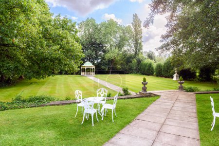 Deserted manicured garden with a gazebo at the end of stone path through a lawn surrounded by trees on a sunny summer day. A metal table and chairs on grass are in foreground. Aston, England, UK.