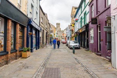 Foto de Caernarfon, Gales, Reino Unido - 10 de julio de 2023: personas caminando por una estrecha calle adoquinada en el casco antiguo amurallado. Caernarfon es una ciudad real, situada en la orilla oriental del estrecho de Menai, frente a la isla de Anglesey. - Imagen libre de derechos