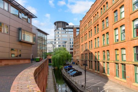 Enger Kanal, gesäumt von einem steinernen Fußweg zwischen einem sanierten Backsteingebäude mit Parkplätzen auf der einen und einem modernen Gebäude auf der anderen Seite an einem sonnigen Sommertag. Manchester, England, Großbritannien.