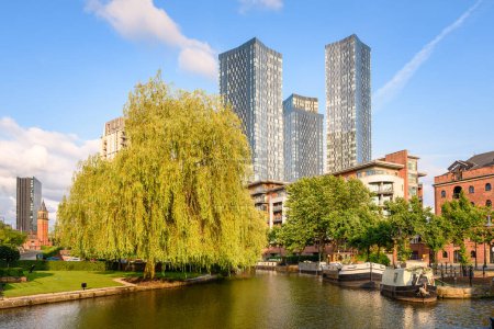 Modern glass residential towers and apartment blocks overlooking a canal with moored barges on a clear summer day. Manchester, England, UK.
