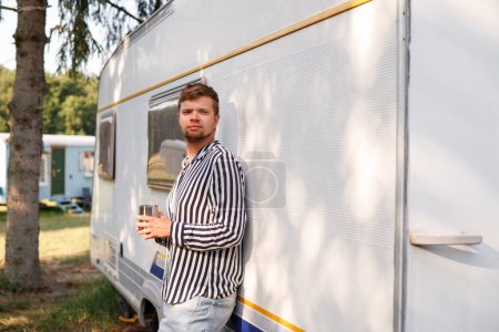 Foto de Retrato de un hombre con una camisa a rayas, sosteniendo una taza, de pie cerca de una casa móvil al aire libre - Imagen libre de derechos