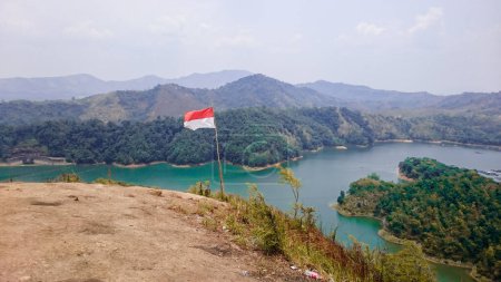 The red and white Indonesian flag stuck on the mountain. You can see other mountain ranges and lakes with green water. There are green and yellow trees, other plants and a cloudy blue sky.