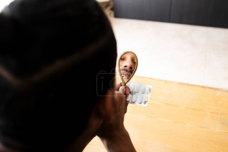 Téléchargez les photos : Latino gars regardant sa dent cassée reflétée dans le miroir et tenant ses médicaments dans sa main. - en image libre de droit