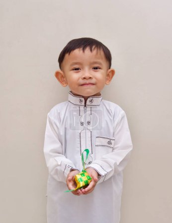 Foto de Un niño lindo de Asia con una camisa larga islámica blanca con un diseño simple, sosteniendo un pequeño Ketupat de color verde y amarillo en su mano, con una expresión facial sonriente - Imagen libre de derechos