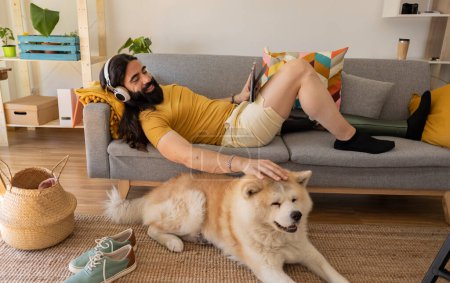 Téléchargez les photos : Homme avec prothèse de jambe sur le canapé écoutant de la musique avec sa tablette, il est à côté de son chien à la maison, - en image libre de droit